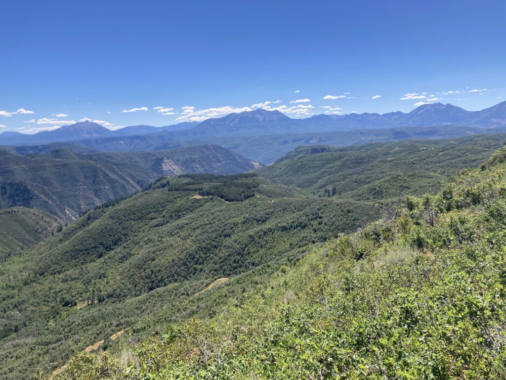 A panoramic view of rolling green hills under a clear blue sky with mountain ranges in the distance.