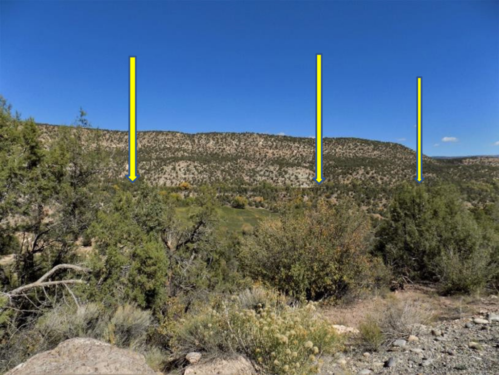 Desert landscape with shrubs, bushes, and rocky hills under a clear blue sky, featuring three yellow bars with arrows pointing downward to indicate the ditch.