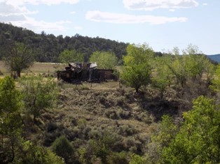 Abandoned building in a rural grassy area surrounded by greenery under a partly cloudy sky.