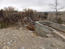 Rusty pipeline and concrete trough in a rocky area with leafless trees in the background.