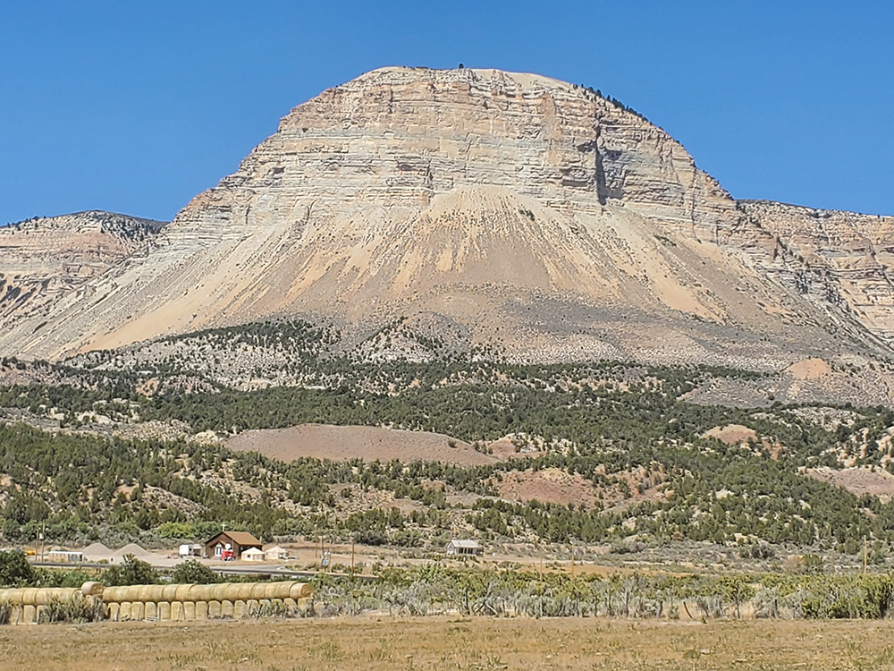 Mountain with layered rock formations, greenery at its base, and a small settlement with a red-roofed building in the foreground. A shadow in the shape of a cross is visible on the mountain face.