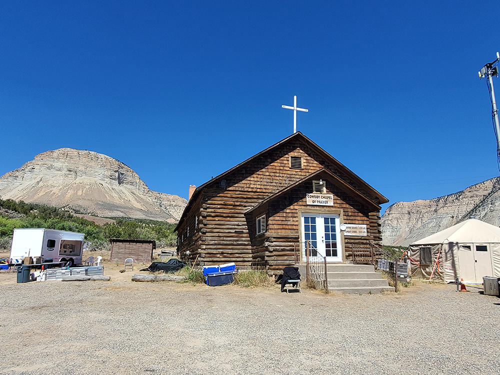 Rustic log chapel in a mountainous area with a trailer, shed, and large tent nearby under a clear blue sky.