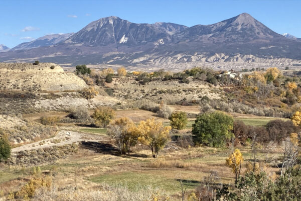 Landscape view of a valley with scattered trees in autumn colors, a path winding through the terrain, and imposing mountains in the background under a clear sky.